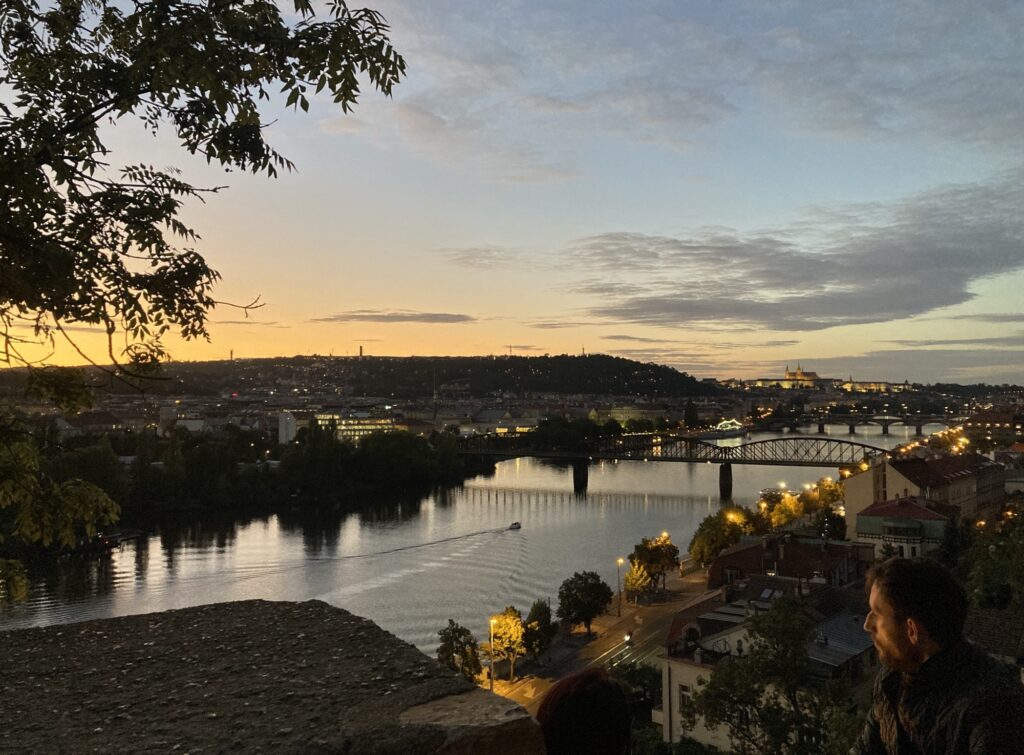 Sonnenuntergang über der Moldau mit Blick auf Bäume, Lichter und ruhige Wasseroberfläche