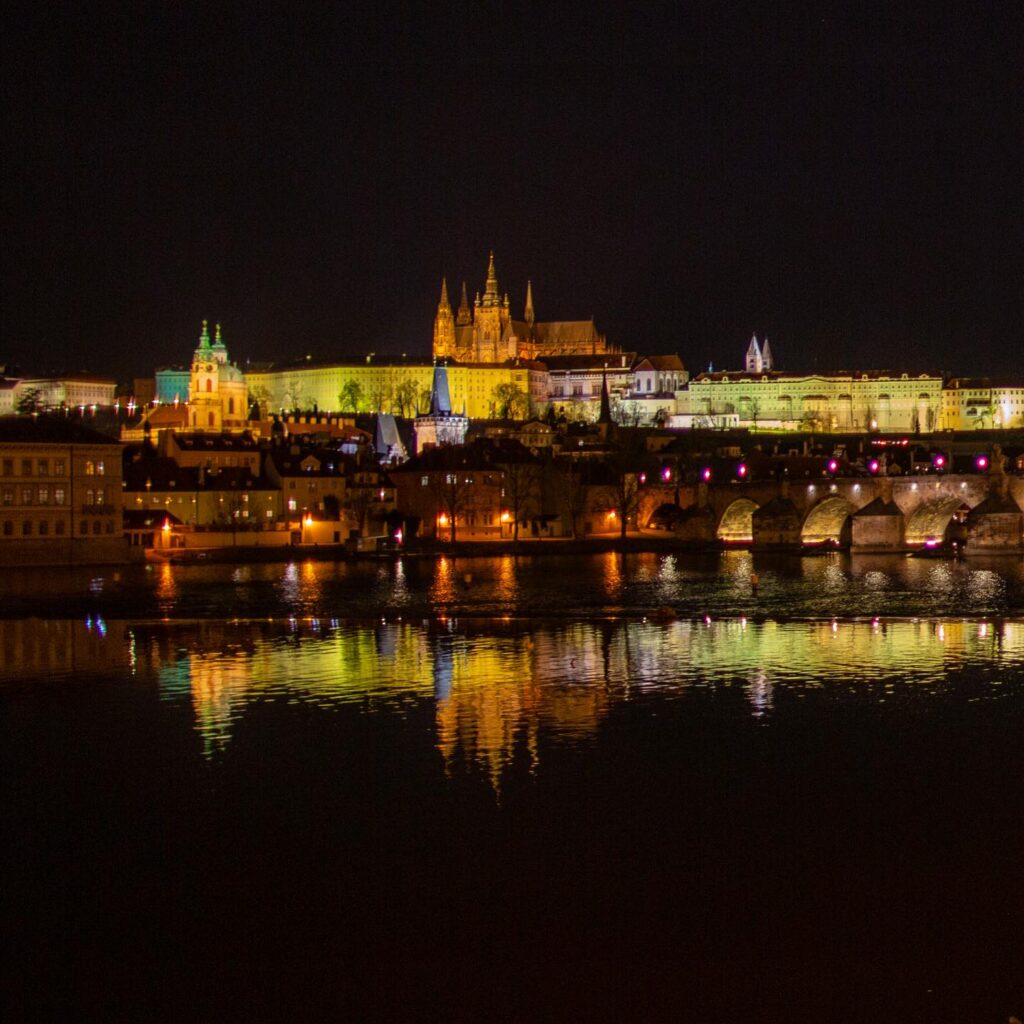Nachtaufnahme mit beleuchteter Prager Burg, Karlsbrücke und den Spiegelungen im ruhigen Wasser der Moldau. Eine ikonische Perspektive auf Prag.