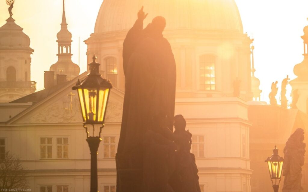 Blick von der Karlsbrücke auf eine Statue mit Laternen und die Prager Altstadt im warmen Morgenlicht