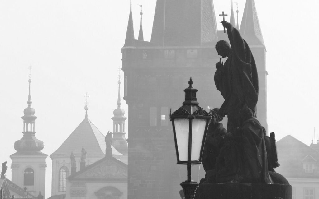 Schwarz-weiße Ansicht einer Statue auf der Karlsbrücke mit Laterne und Brückenturm im Hintergrund