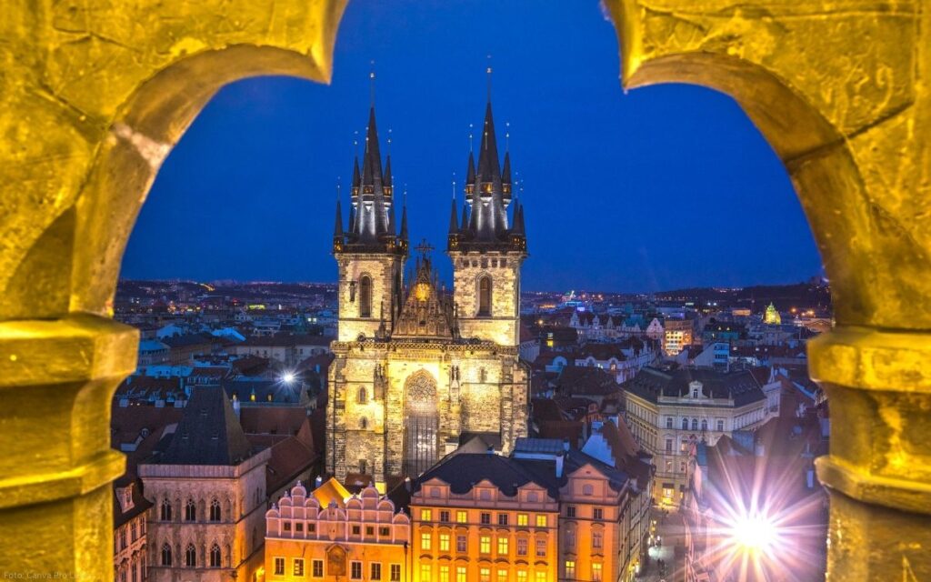 Blick auf die Prager Altstadt mit der Teynkirche bei Nacht, eingerahmt von einem beleuchteten gotischen Fenster am Altstädter Ring