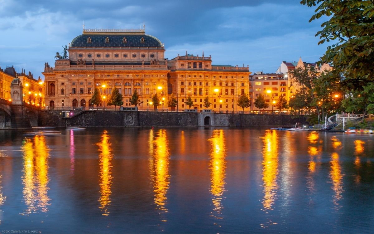 Nationaltheater in Prag bei Abenddämmerung mit beleuchteter Fassade und Spiegelungen in der Moldau
