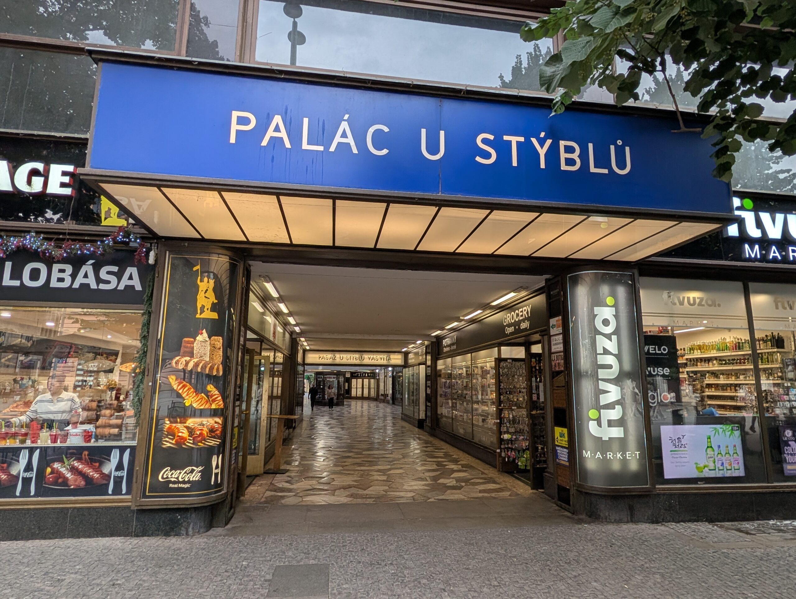 Eingang zum Palast U Stýblů in Prag mit blauem Schriftzug, Passage und angrenzenden Geschäften