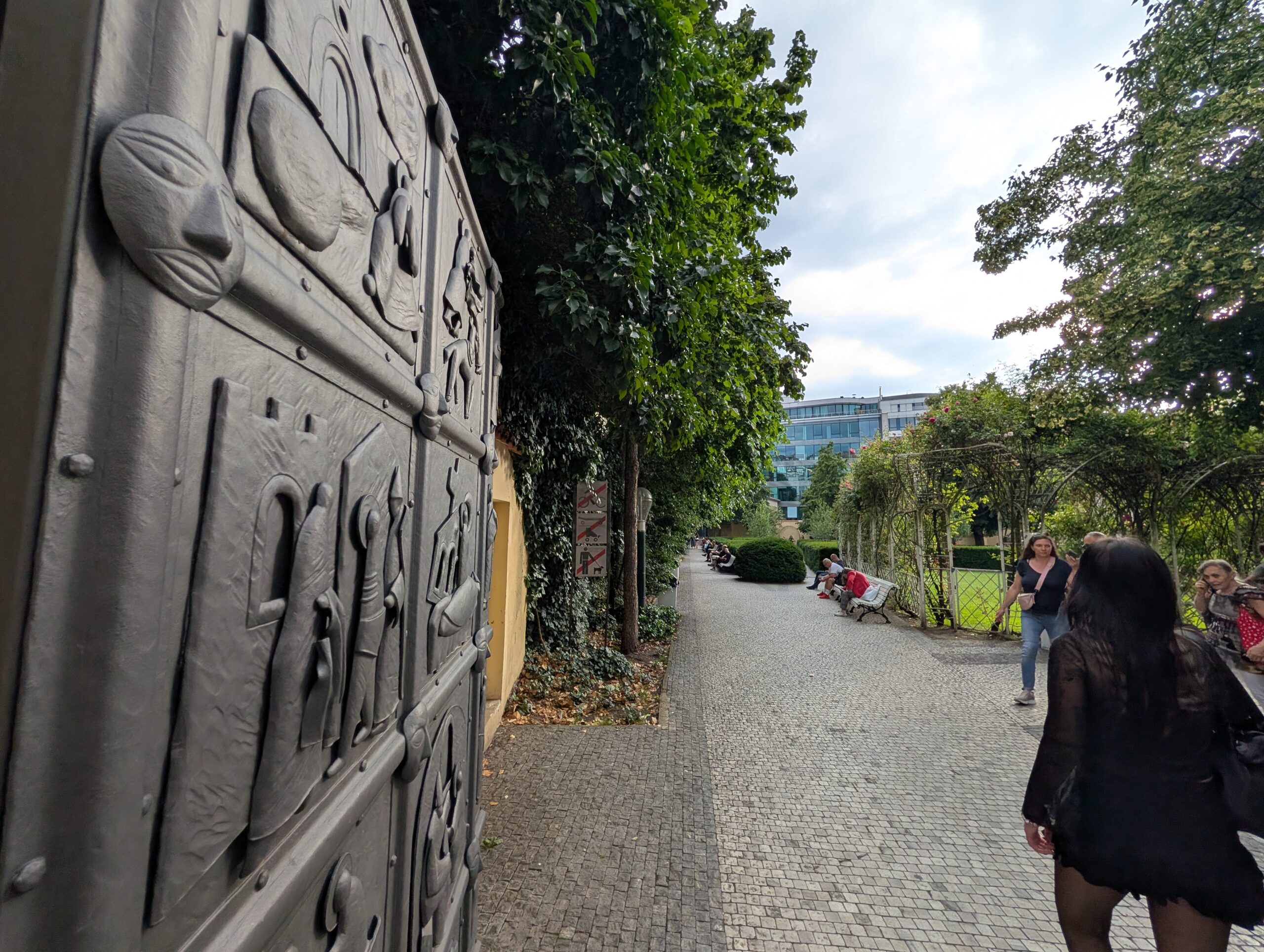 Verziertes Metalltor am Eingang zum Franziskanergarten in Prag mit gepflastertem Weg, Bäumen und Besuchern