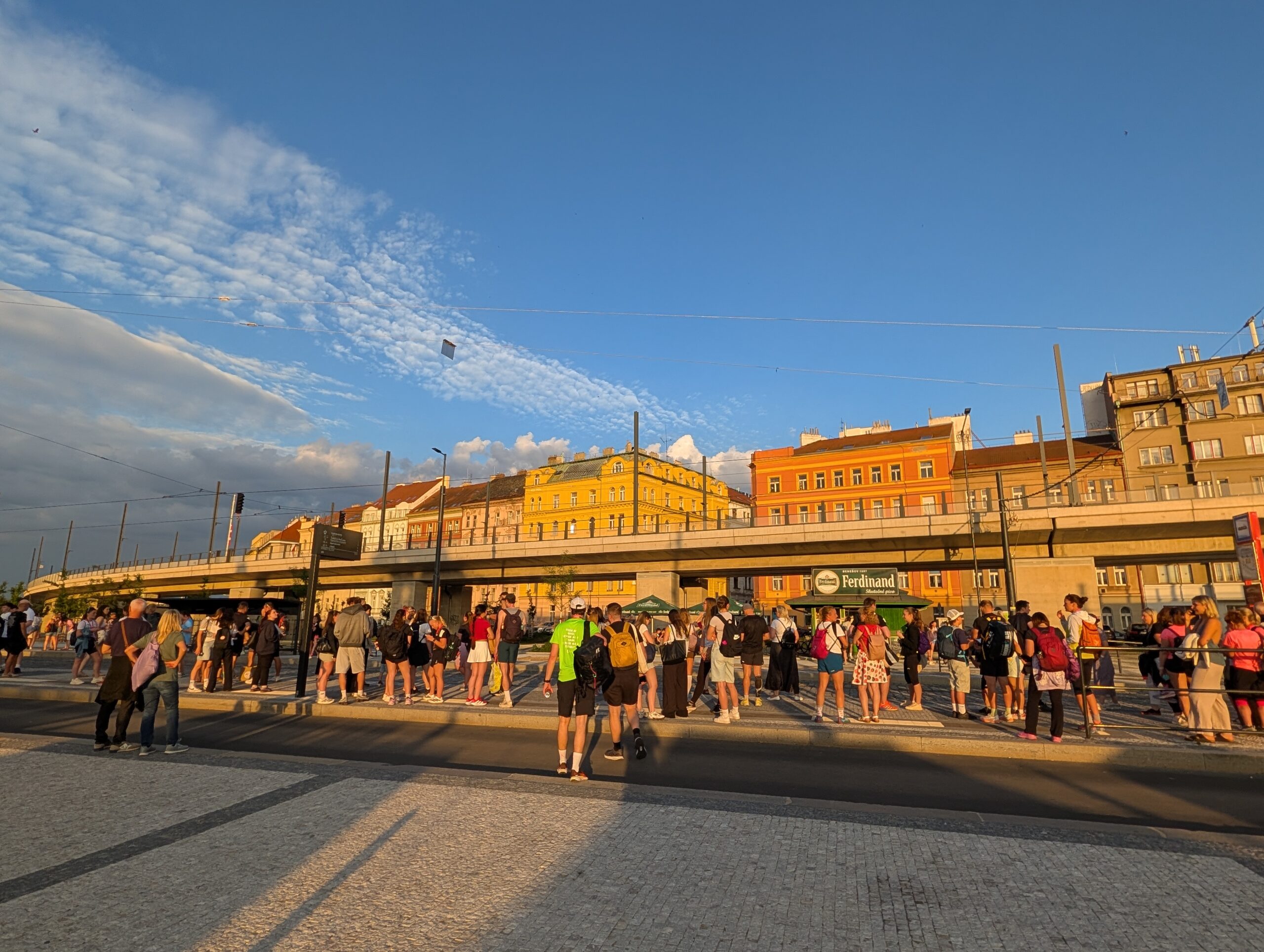 Besucher auf dem Vorplatz des Messegeländes Prag mit Haltestelle, Oberleitungen und Häuserzeile im Abendlicht