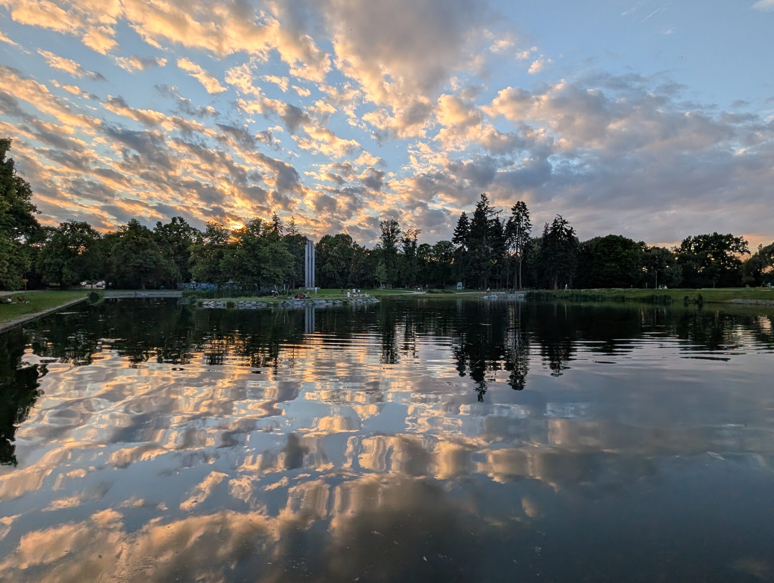 Parkteich bei Sonnenuntergang mit spiegelnden Wolken, Bäumen und moderner Säulenskulptur