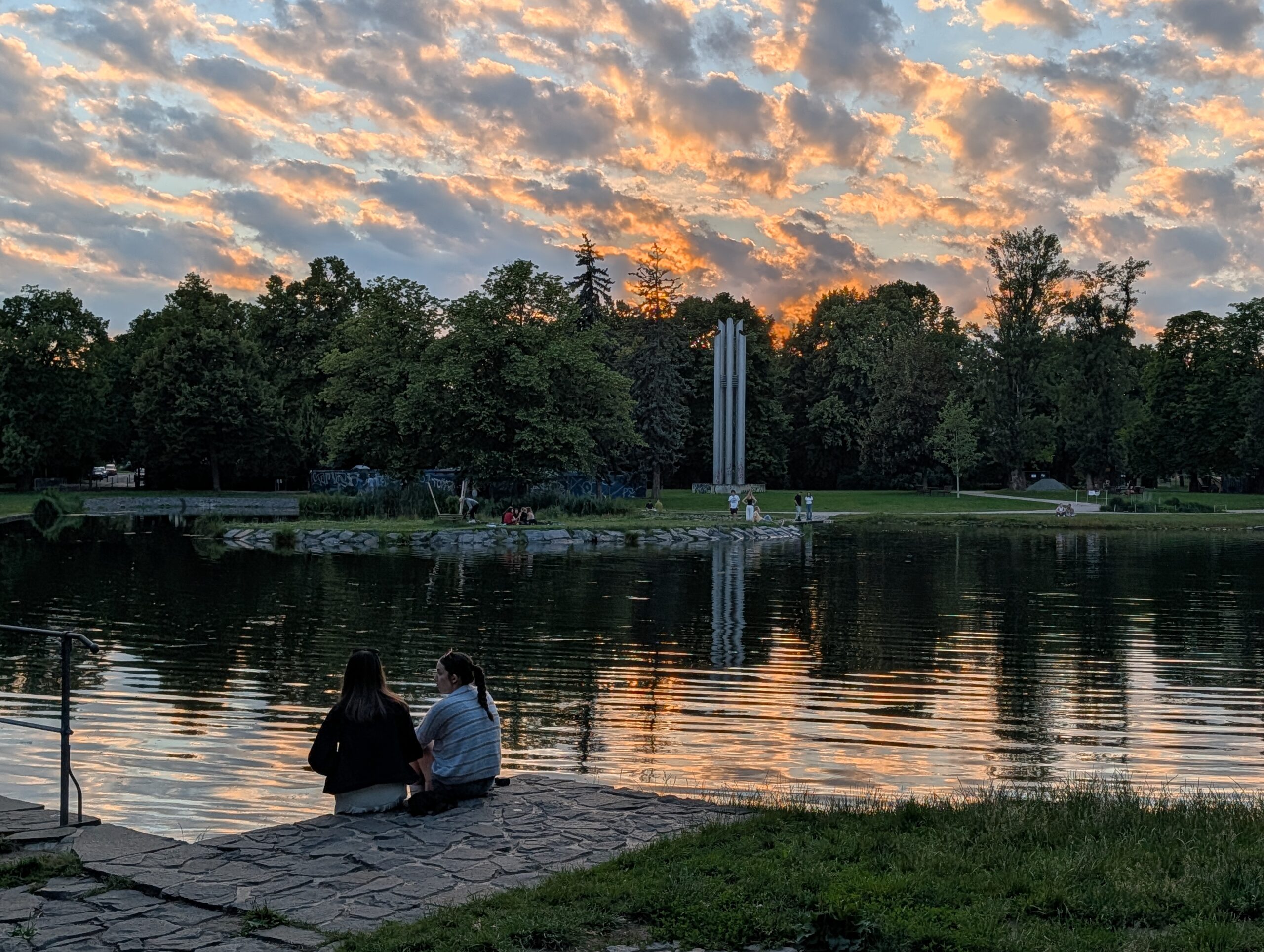 Parkteich bei Sonnenuntergang mit zwei Personen am Ufer, Bäumen und moderner Säulenskulptur