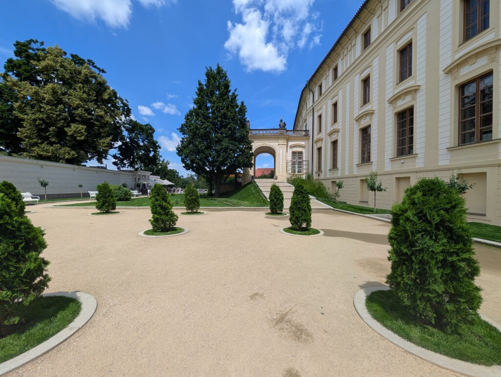 Garten der Prager Burg mit Kiesfläche, geschnittenen Sträuchern, historischem Gebäudeflügel und Torbogen