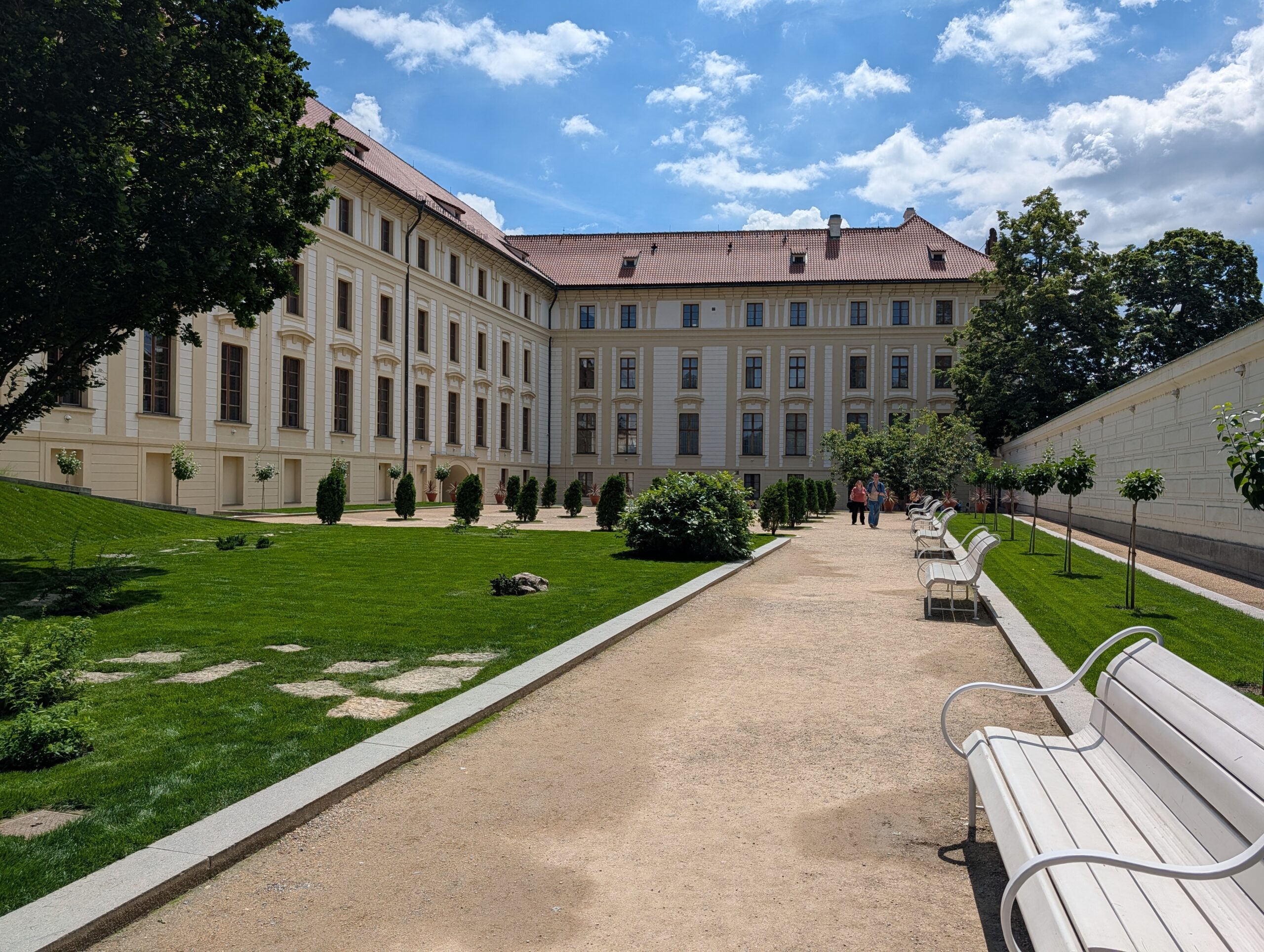 Hartig-Garten an der Prager Burg mit Kiesweg, Rasenflächen, weißen Sitzbänken und historischem Gebäude