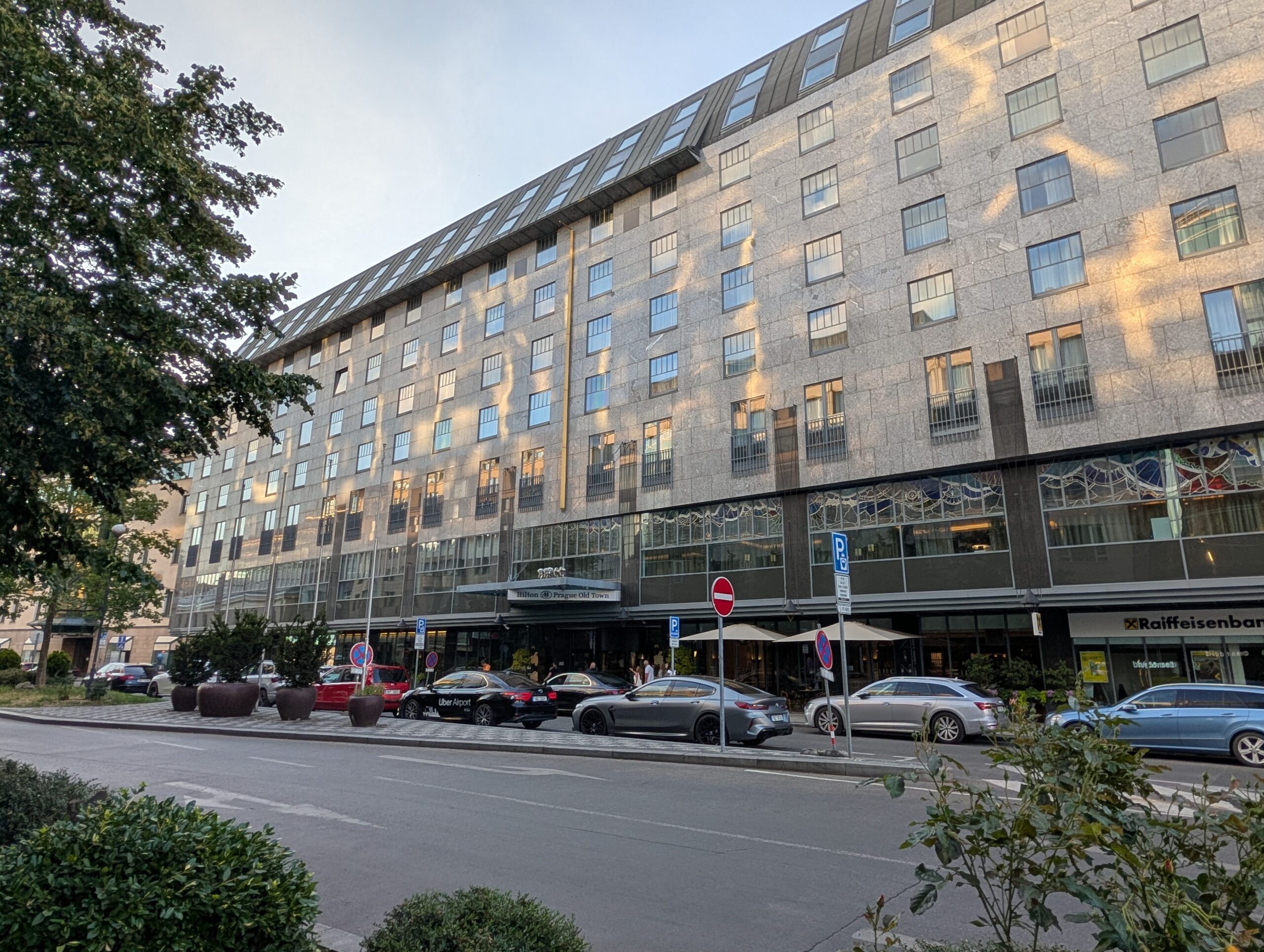 Langgestreckte Fassade des Hilton Prague Old Town mit Hoteleingang, parkenden Autos und Grünflächen