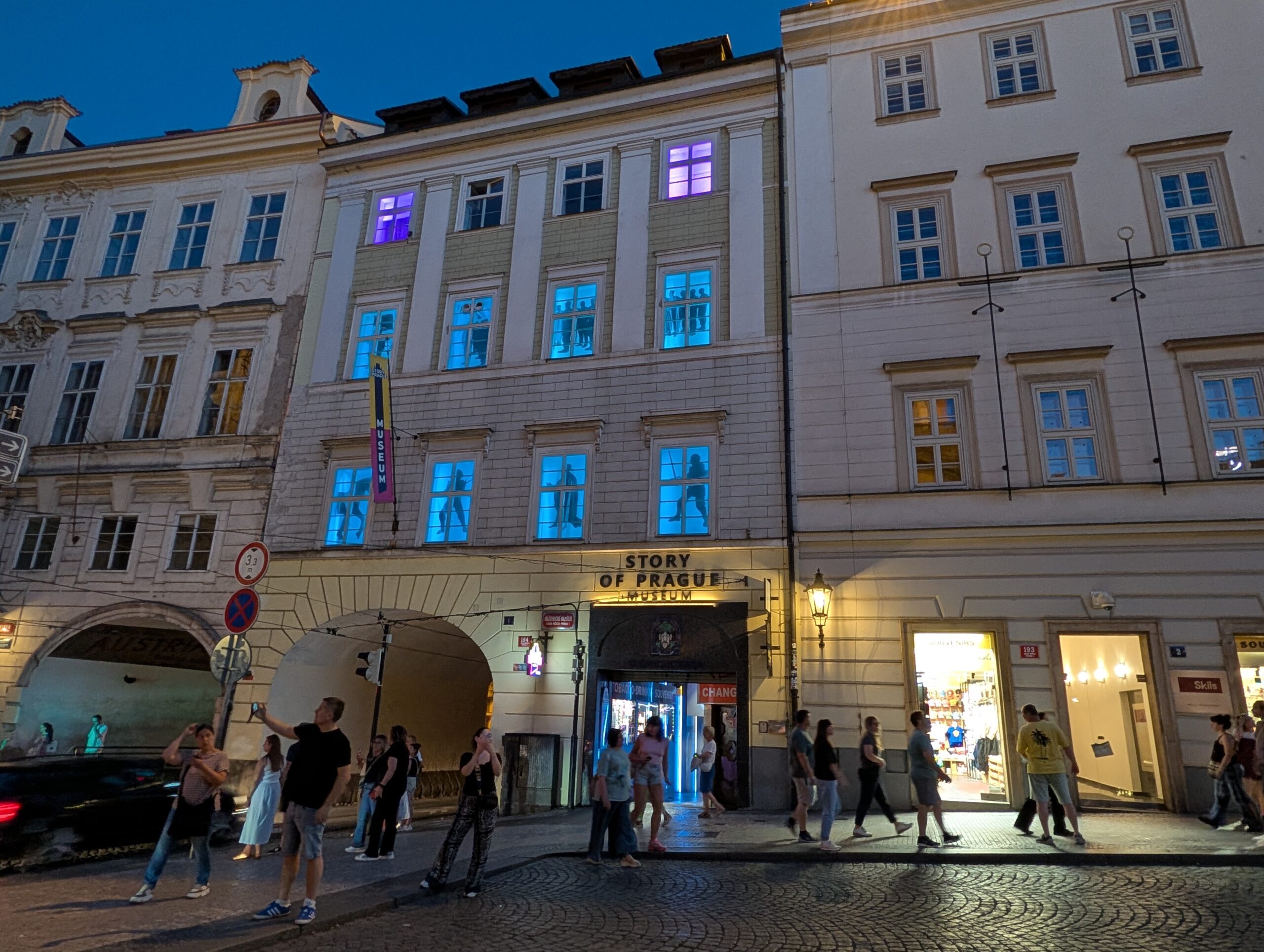 Beleuchteter Eingang zum Story of Prague Museum in historischer Häuserzeile mit Passanten bei Nacht