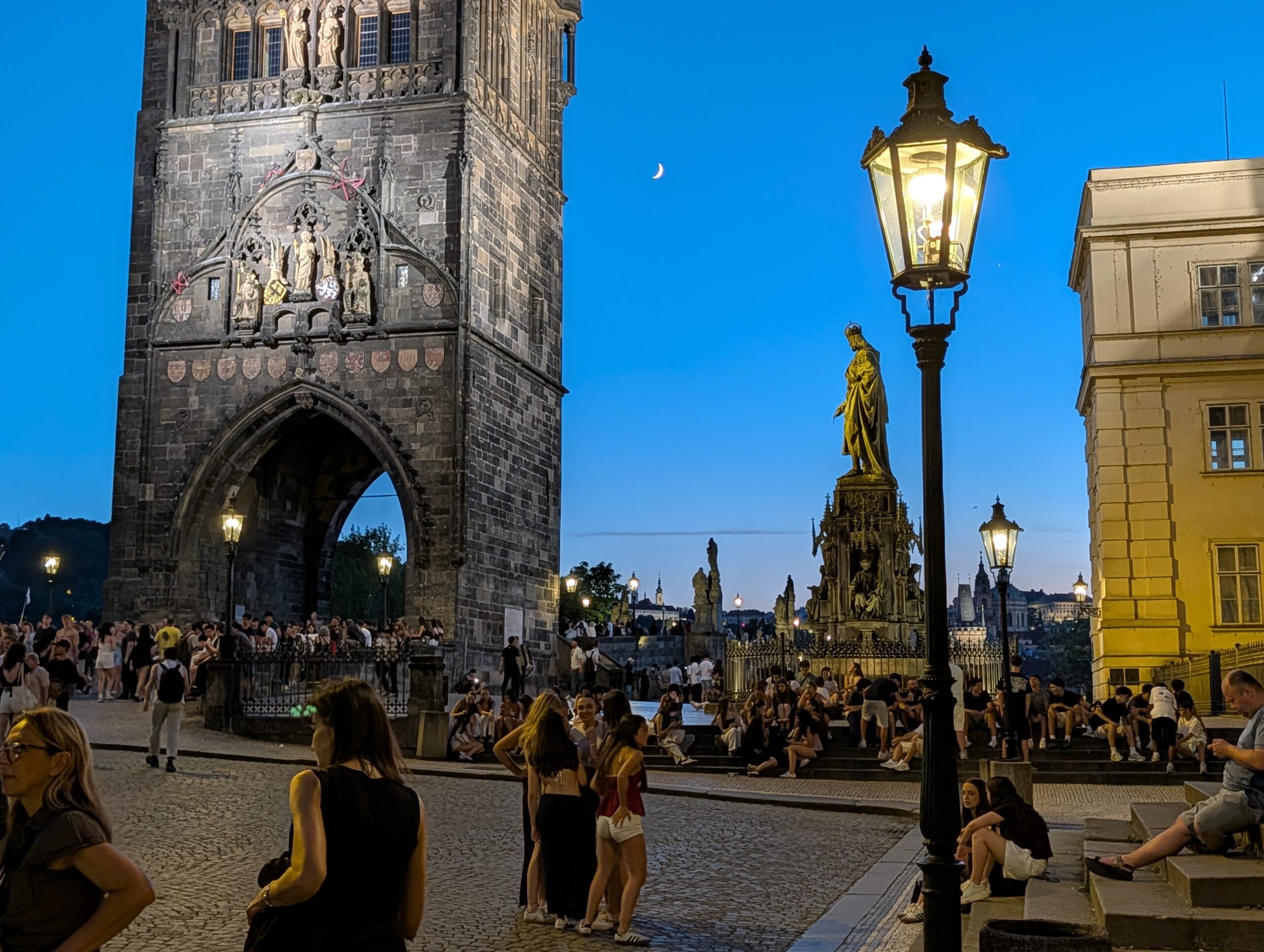 Belebter Kreuzherrenplatz mit Altstädter Brückenturm, Karls-IV.-Denkmal, Straßenlaternen und Mondsichel am Abend