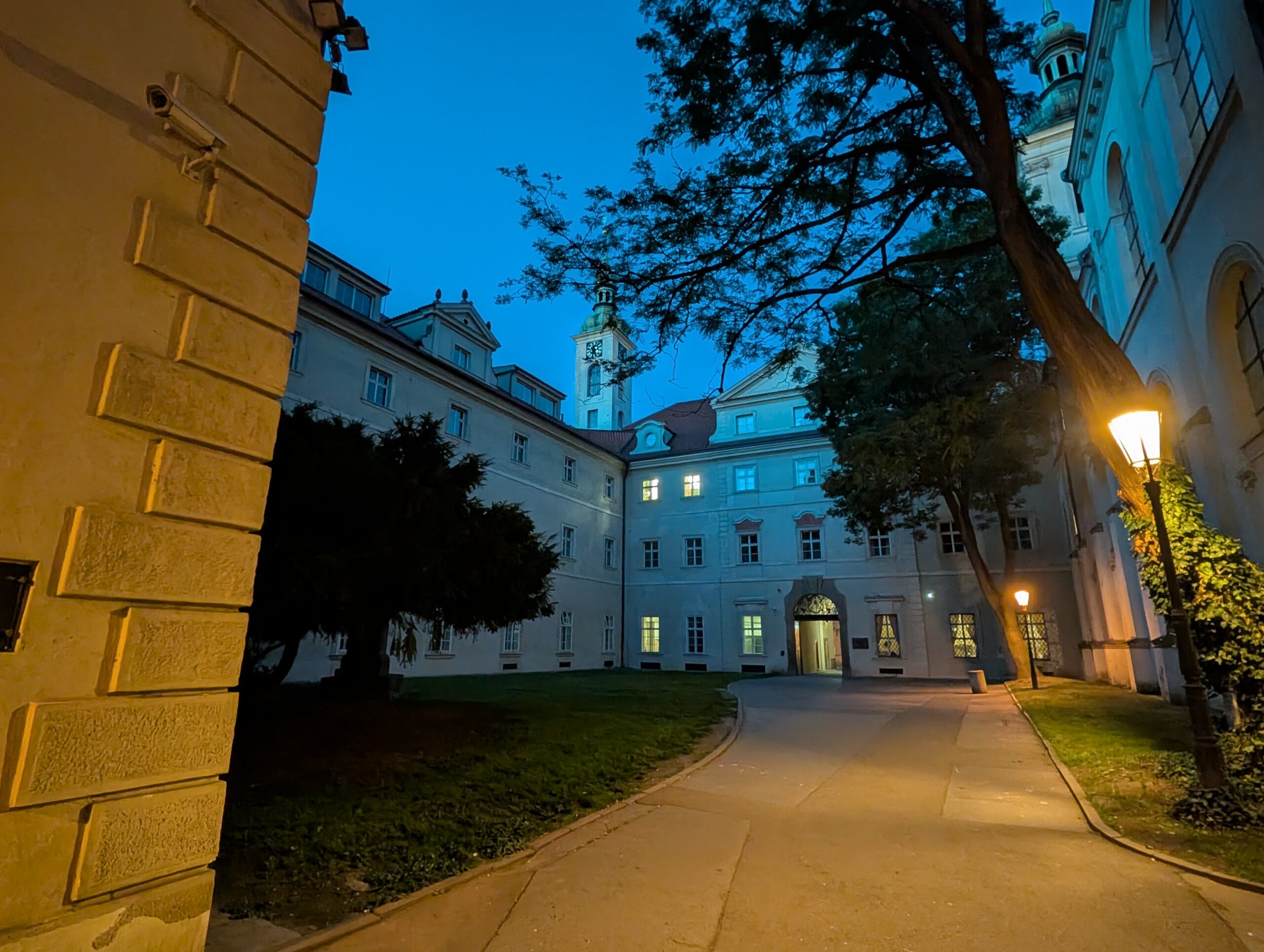 Beleuchteter Innenhof des Klementinums mit geschwungenem Weg, Bäumen, historischen Fassaden und Turm bei Nacht