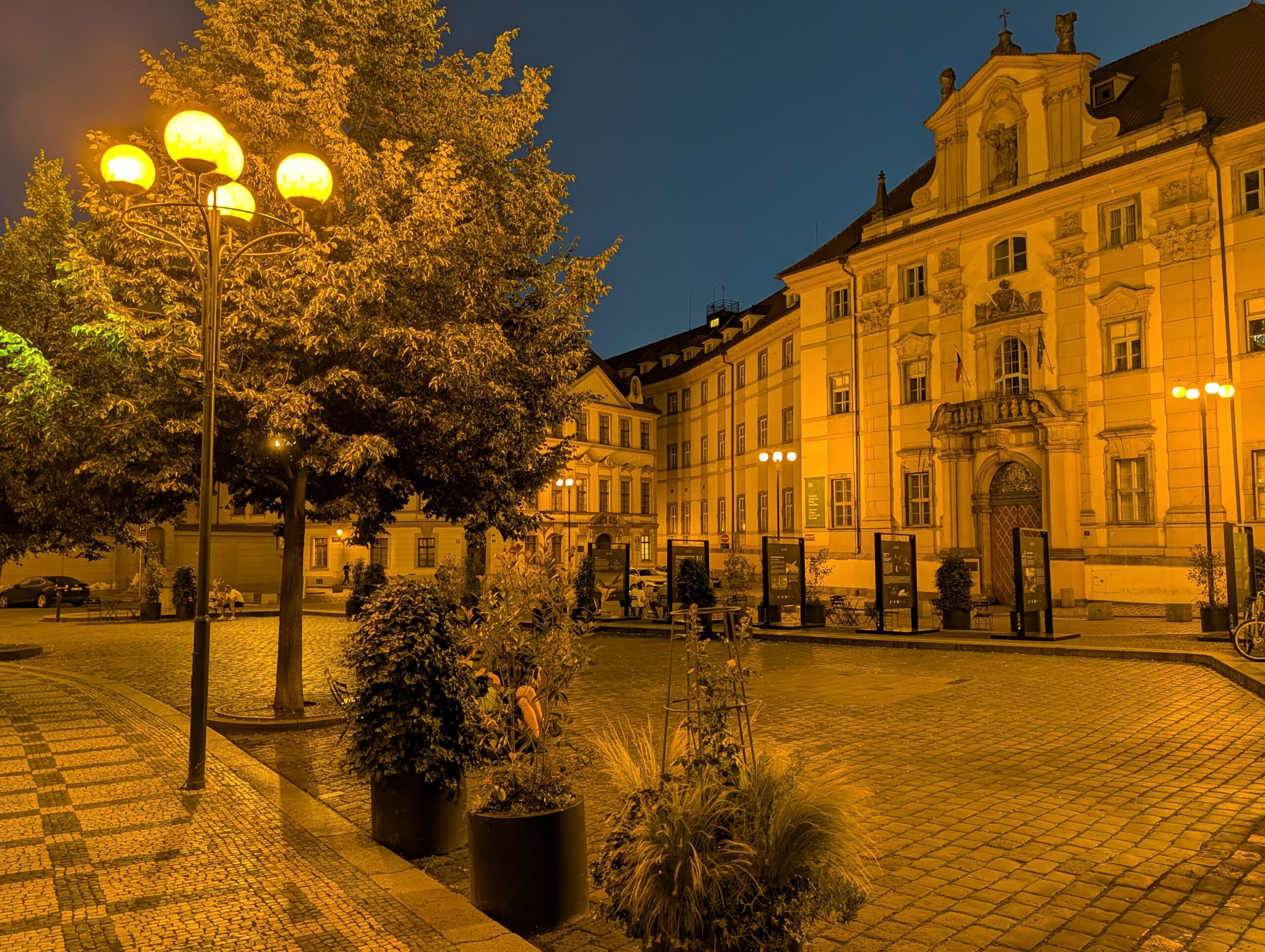 Marienplatz in Prag bei Nacht mit historischen Gebäuden, Straßenlaternen, Bäumen und Eingang zum Klementinum