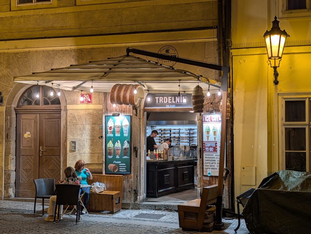 Beleuchteter Verkaufsstand für Baumstriezel mit Sitzplätzen und historischer Straßenlaterne in Prag bei Nacht