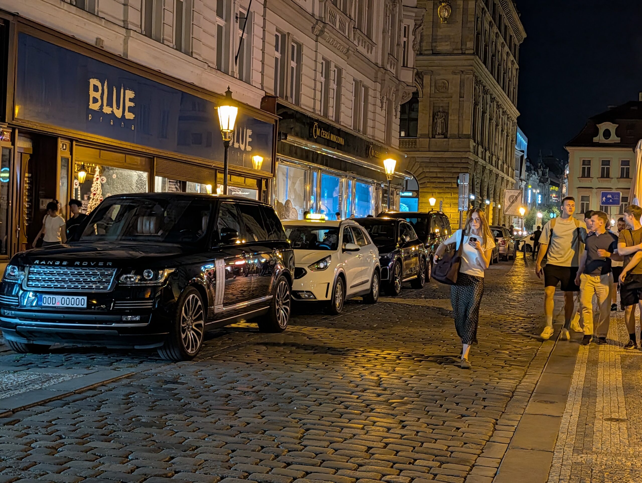 Beleuchteter Eingang von Blue Praha mit parkenden Autos und Passanten in der Prager Altstadt bei Nacht