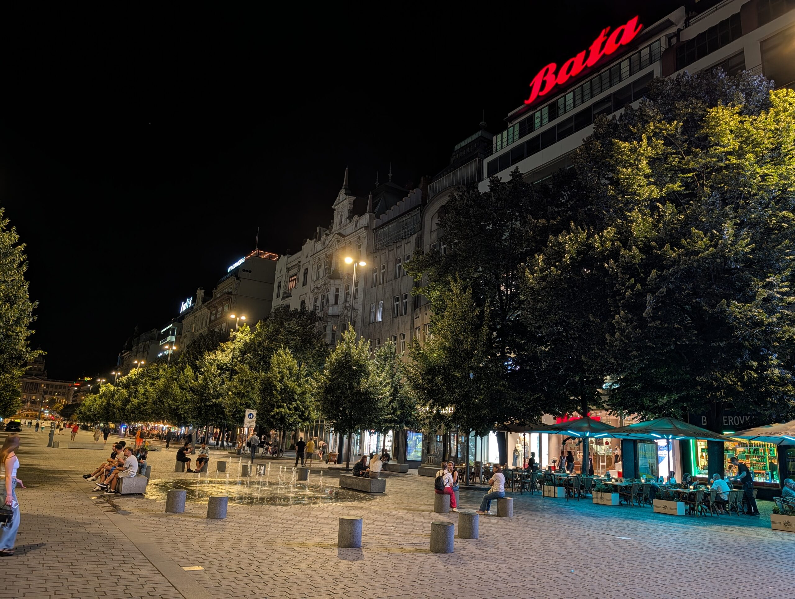 Wenzelsplatz in Prag bei Nacht mit Straßencafés, Bäumen und roter Baťa-Leuchtreklame