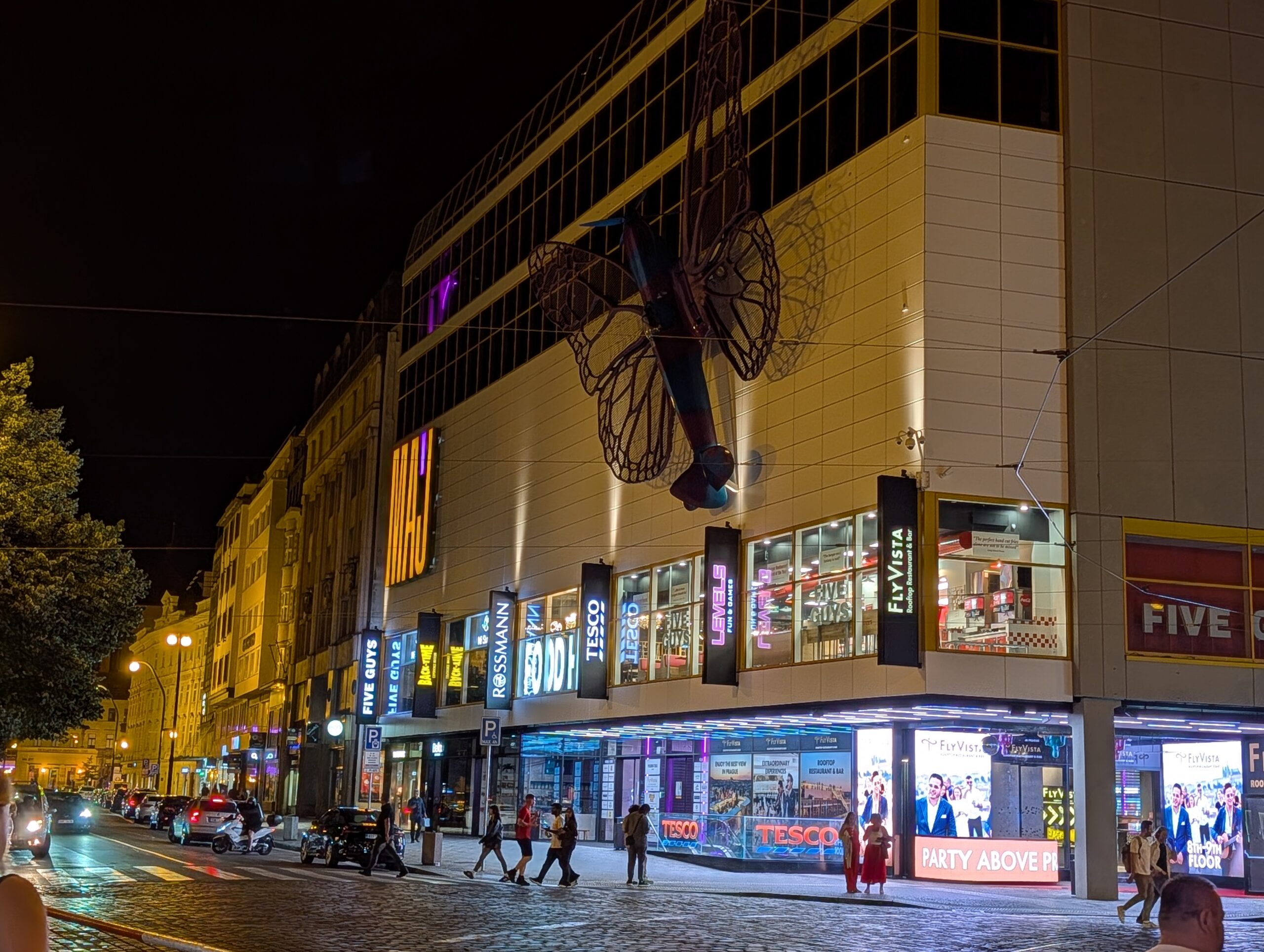 Beleuchtetes Einkaufszentrum MY Národní mit FlyVista, Geschäften und großer Schmetterlingsskulptur an der Fassade bei Nacht in Prag.
