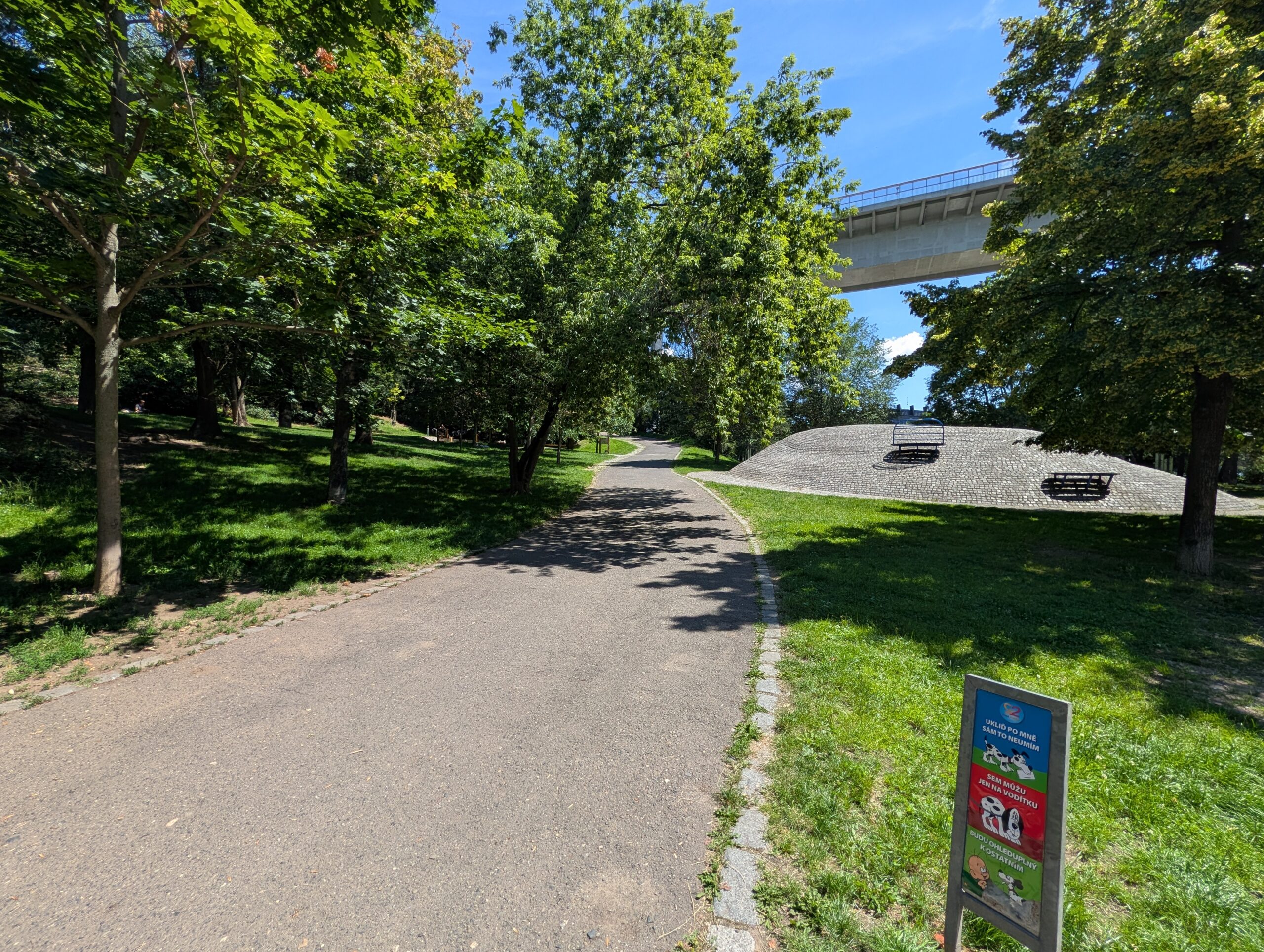 Spazierweg im Park Folimanka unter der Nusle-Brücke in Prag mit Bäumen und Grünflächen