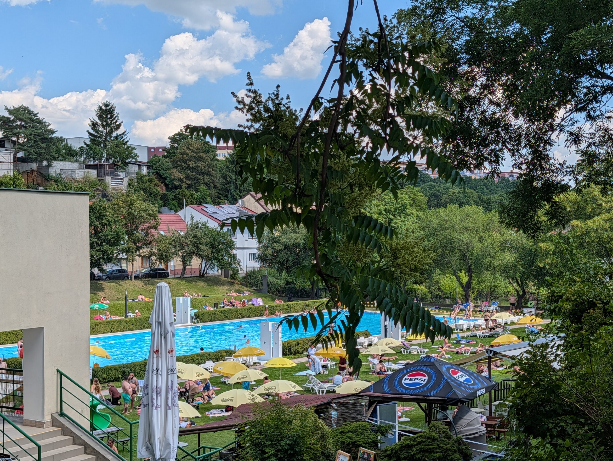 Blick über das Freibad Stírka (Koupaliště Stírka) in Prag mit großem Schwimmbecken, Liegewiesen, Sonnenschirmen und zahlreichen Badegästen.