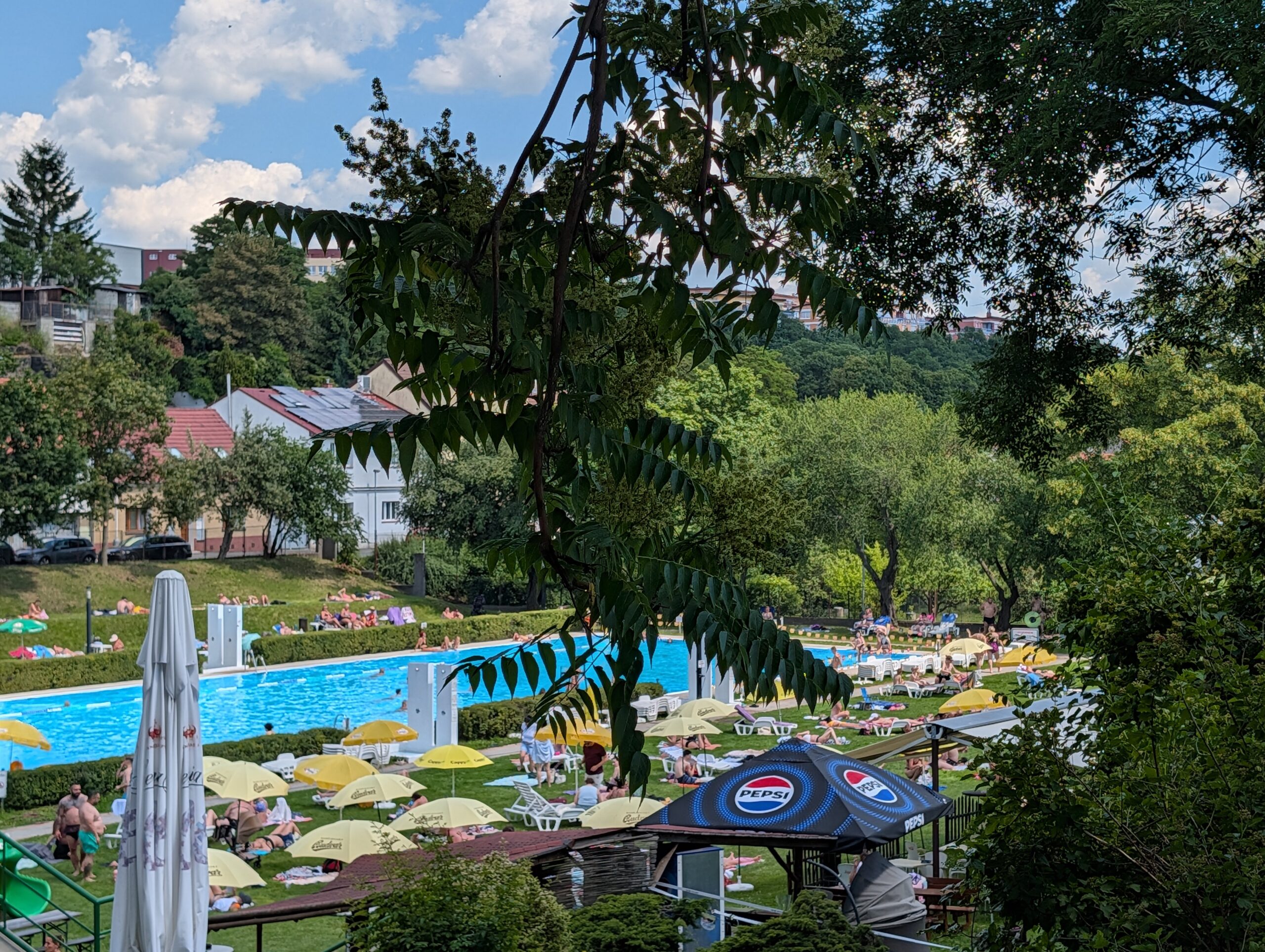 Blick über das Freibad Stírka (Koupaliště Stírka) in Prag mit großem Schwimmbecken, Liegewiesen, Sonnenschirmen und zahlreichen Badegästen.