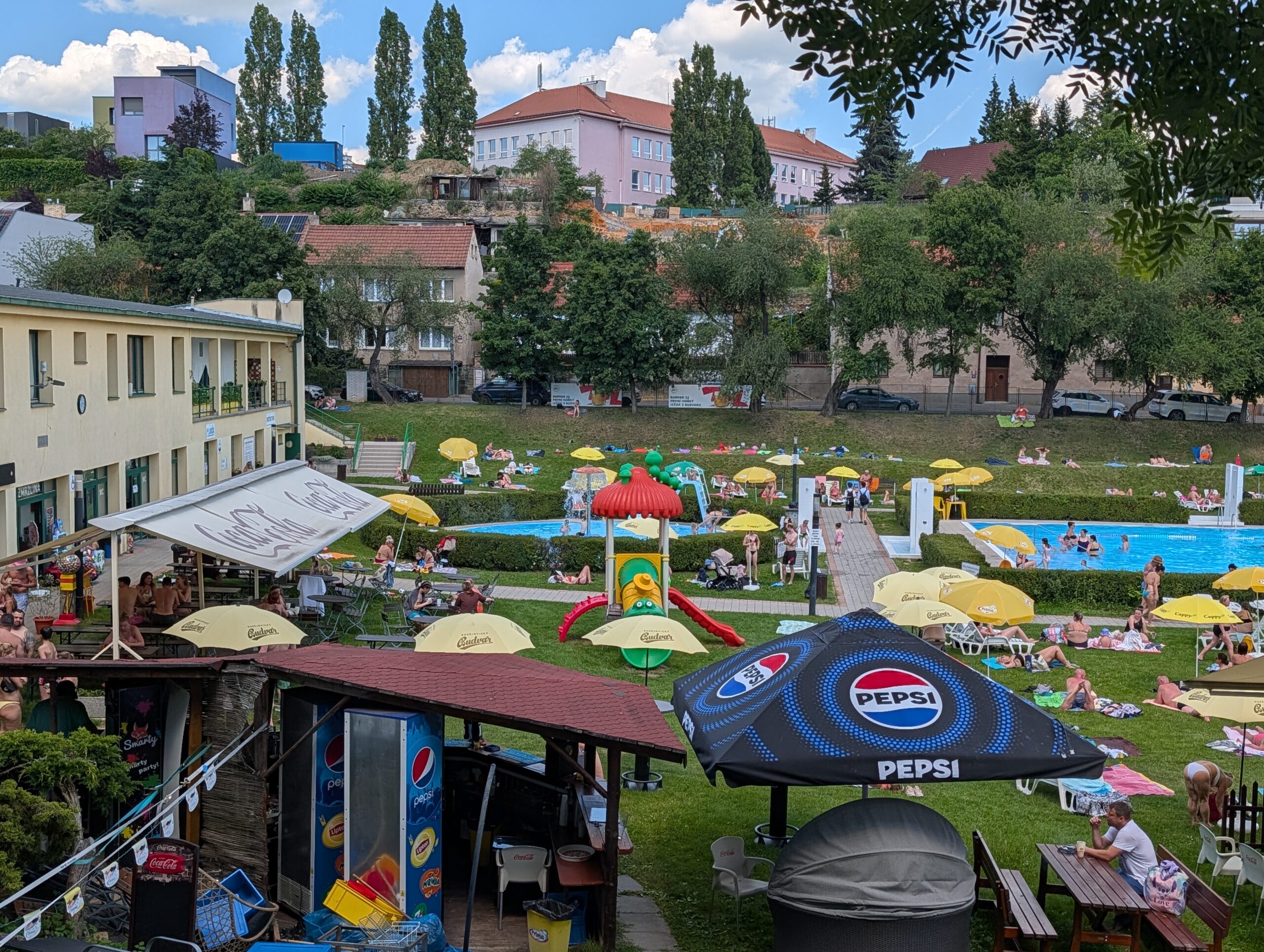 Blick über das Freibad Stírka (Koupaliště Stírka) in Prag mit Liegewiese, Schwimmbecken, Sonnenschirmen und zahlreichen Badegästen.