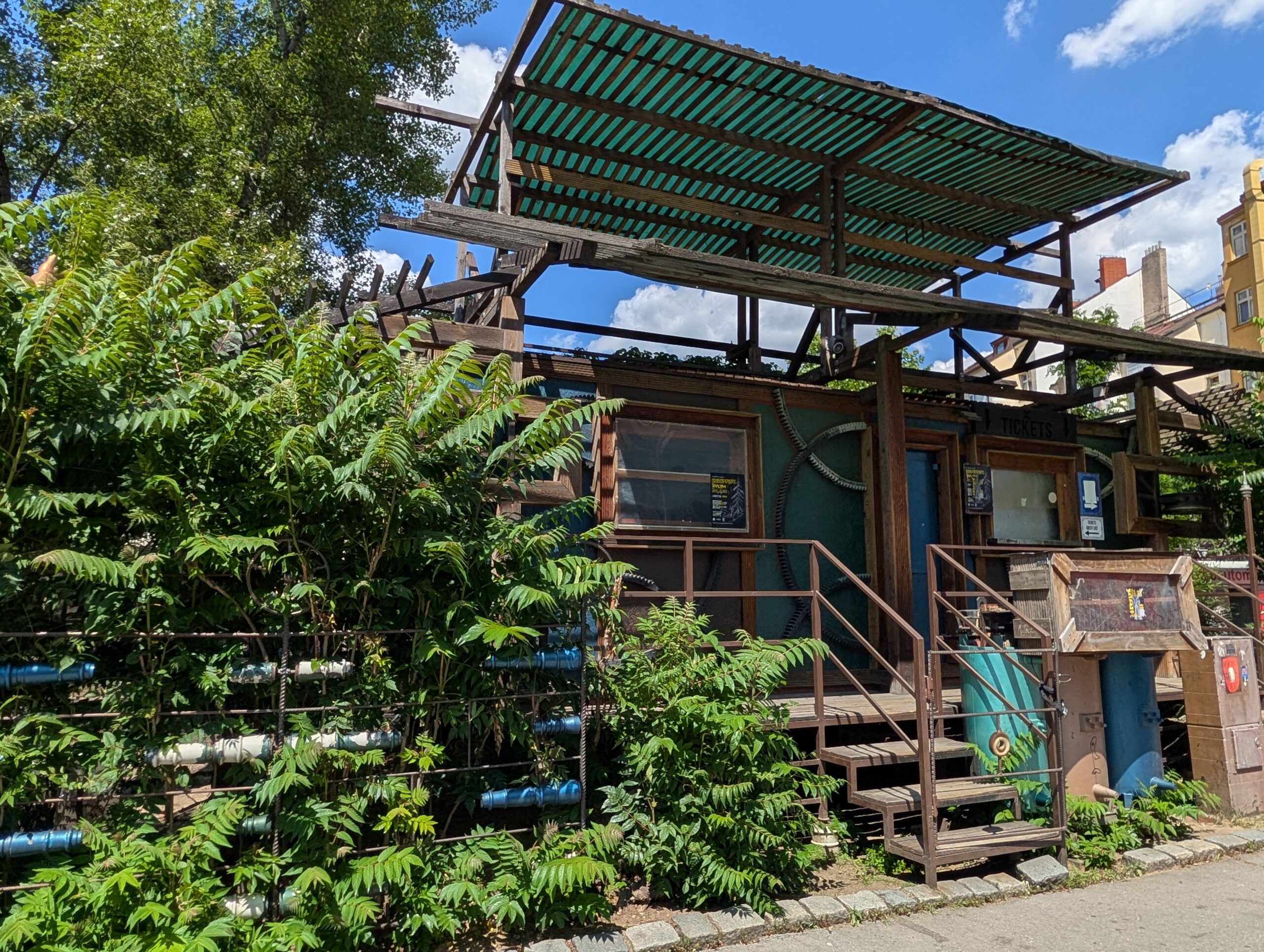 Rustikale Außenbar im Beer Garden des Cross Club in Prag-Holešovice mit Metallkonstruktionen, Holzdetails und üppiger Begrünung.