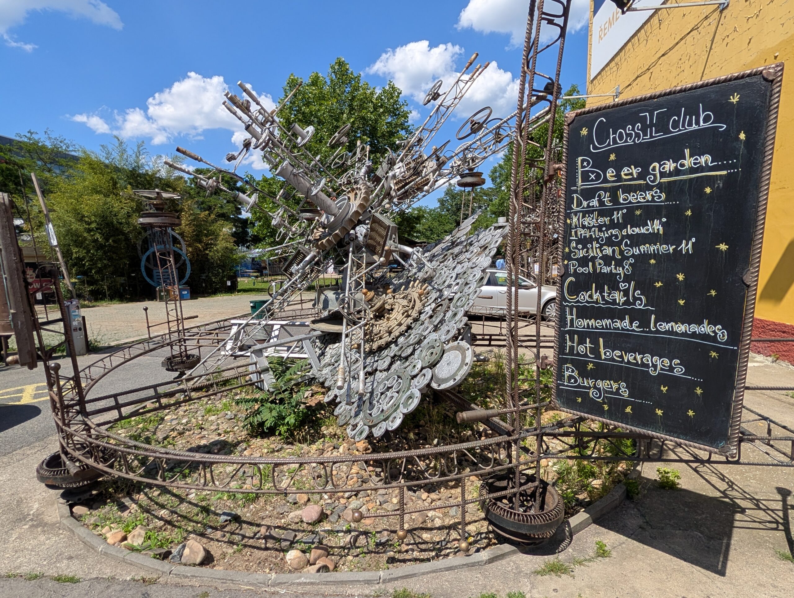 Beer Garden des Cross Club in Prag-Holešovice mit Steampunk-Kunstwerken, dekorativen Metallkonstruktionen und Infotafel im Außenbereich.