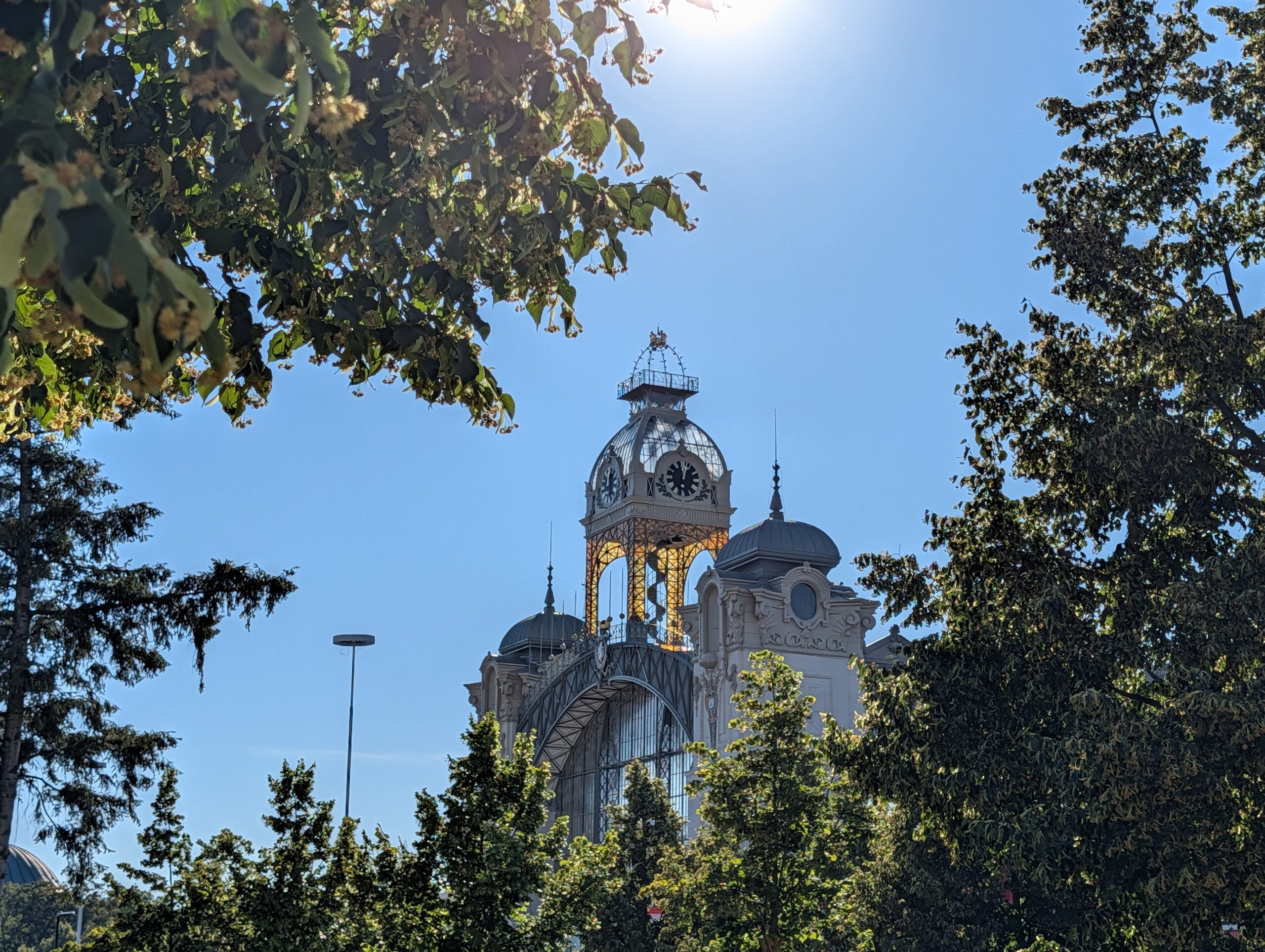 Uhrturm des Industriepalasts (Průmyslový palác) auf dem Prager Messegelände (Výstaviště Praha), teilweise von Bäumen umgeben.