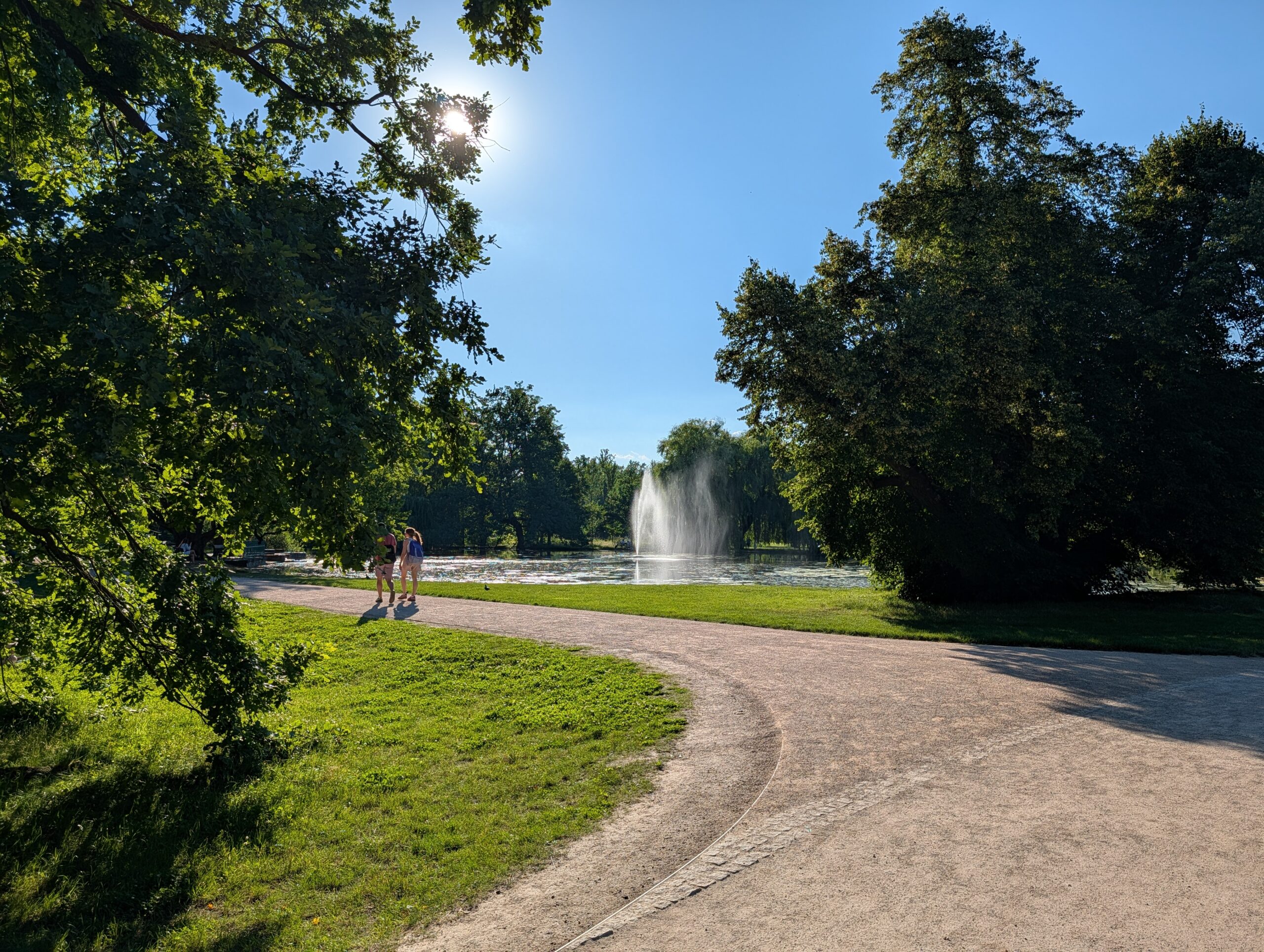 Geschwungener Spazierweg mit Blick auf einen Teich und eine Wasserfontäne im Königlichen Wildpark Stromovka (Královská obora Stromovka) in Prag.