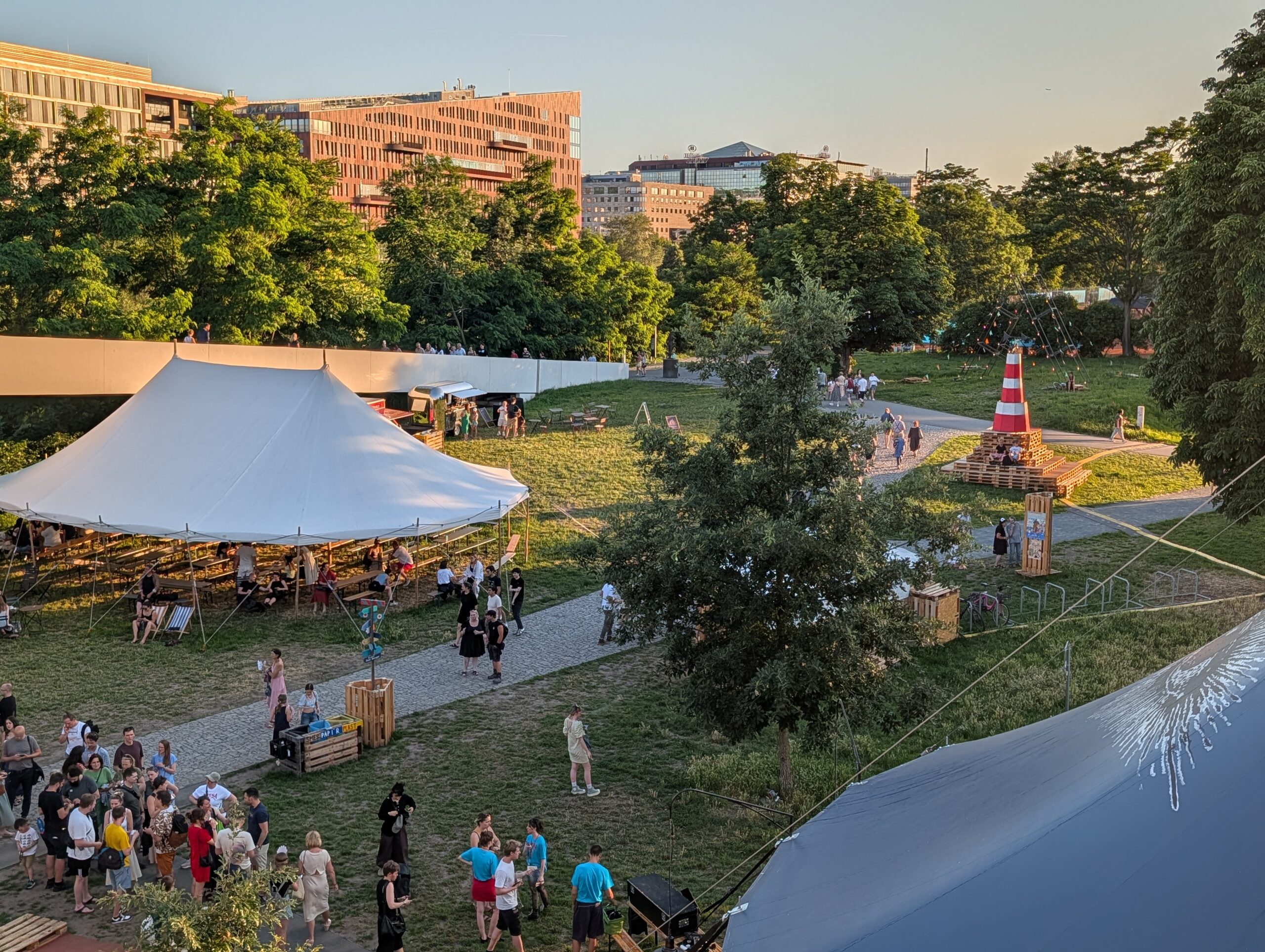 Festivalgelände des ARENA Festivals auf der Insel Štvanice in Prag mit Zirkuszelt, Besuchern und der HolKa-Fußgängerbrücke im Hintergrund.