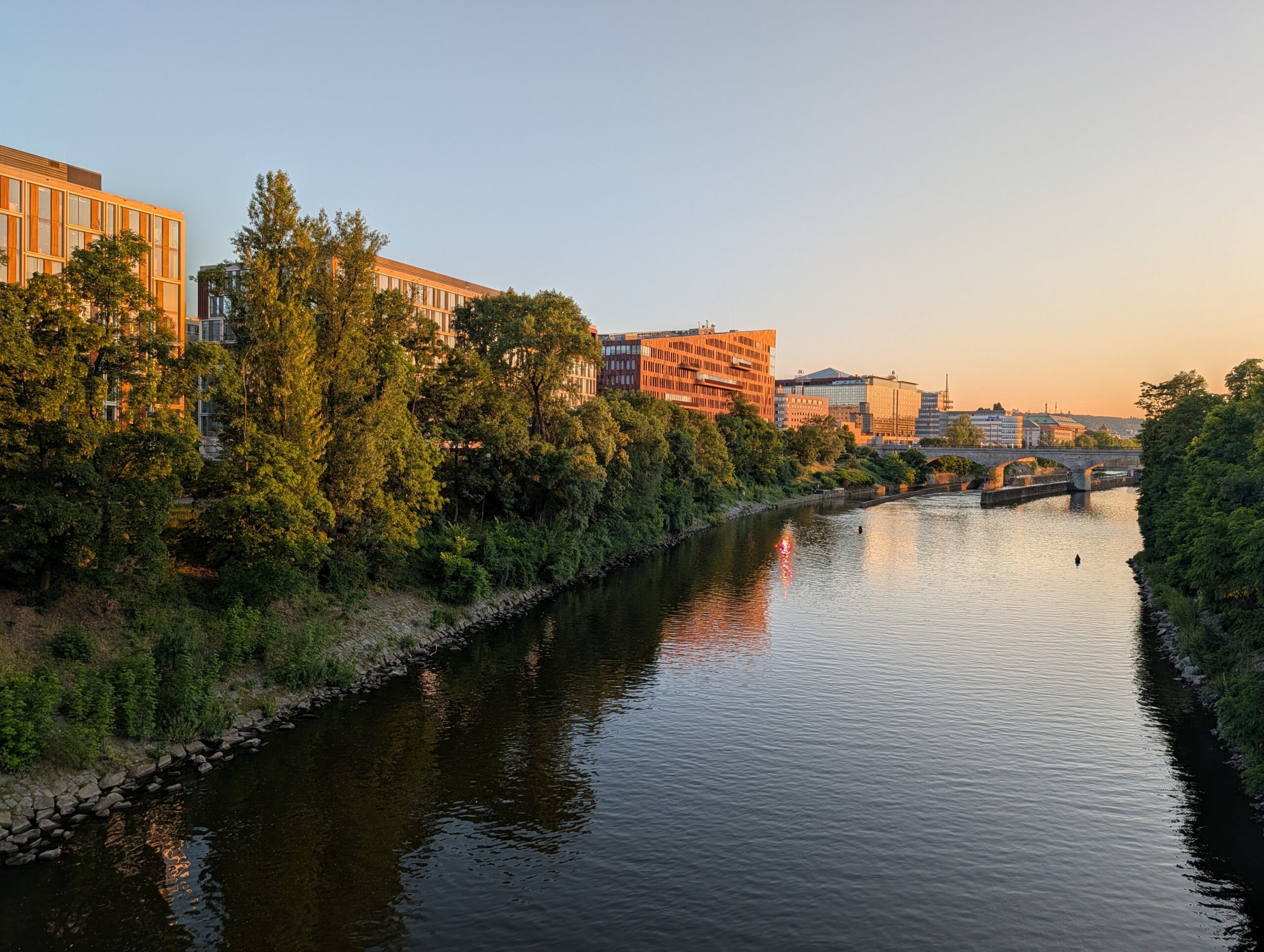 Blick von Karlín über die Moldau zur Insel Štvanice in Prag bei Sonnenuntergang mit moderner Uferbebauung und ruhigem Fluss.