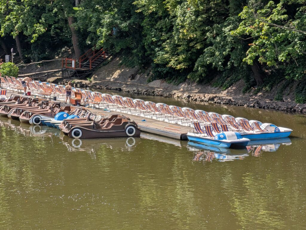 Mehrere Tretboote an einem Steg am bewaldeten Ufer der Moldau in Prag