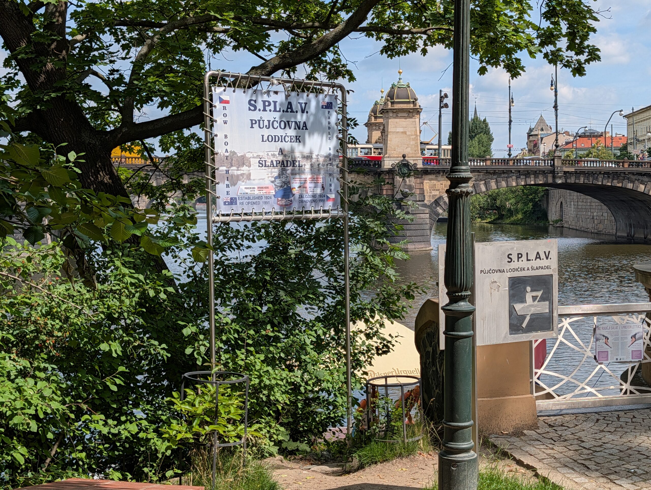 Zugang zum Bootsverleih S.P.L.A.V. an der Moldau mit Brücke und Straßenbahn im Hintergrund