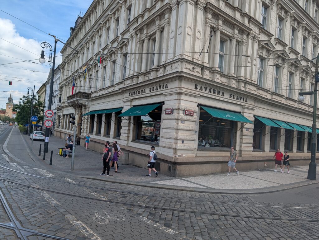 Café Slavia an einer Prager Straßenecke mit grünen Markisen, Fußgängern und Straßenbahngleisen