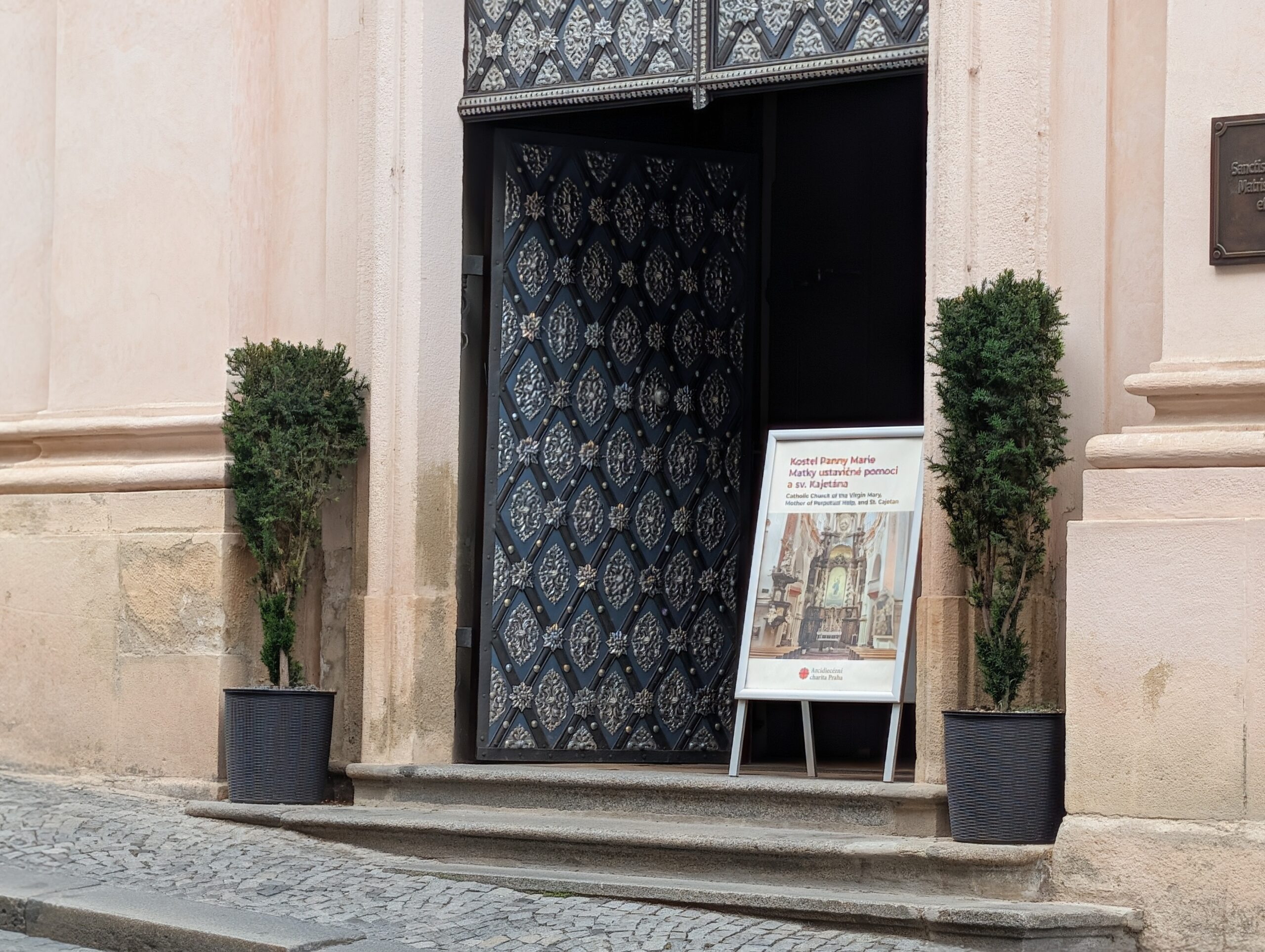 Geöffnetes verziertes Kirchenportal mit Informationstafel und zwei Pflanzen in Prag