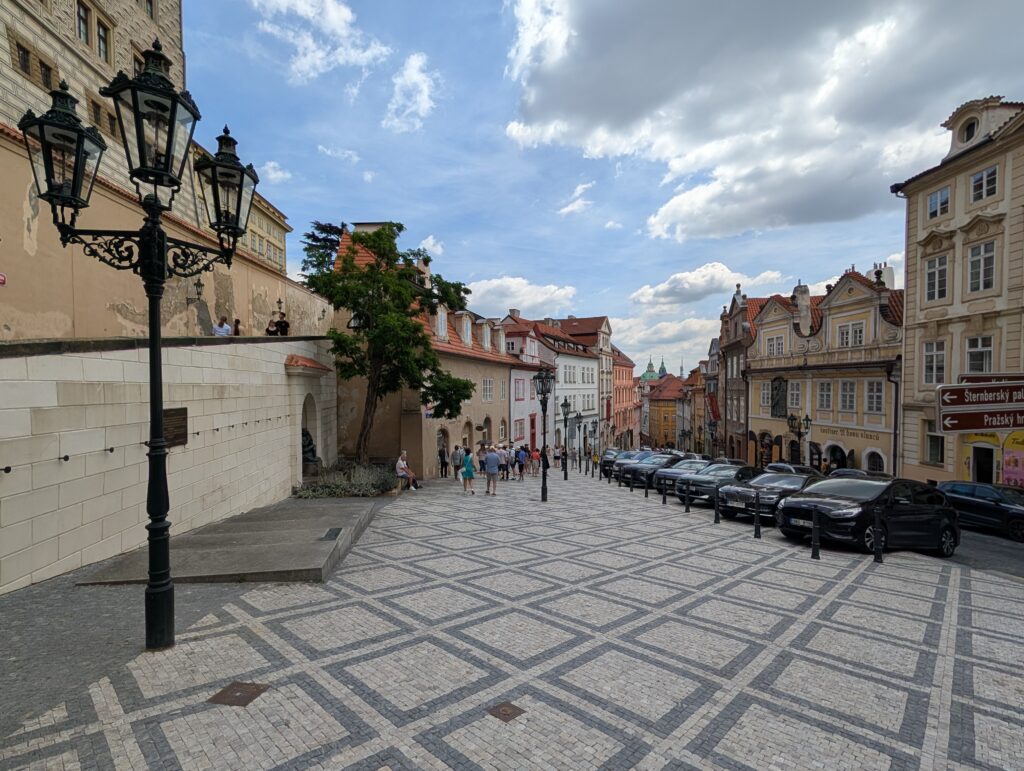 Die Straße Ke Hradu auf dem Prager Hradschin verläuft zwischen historischen Gebäuden in Richtung Prager Burg. Geometrisch gemustertes Pflaster, reich verzierte Straßenlaternen, geparkte Fahrzeuge und mehrere Personen prägen die ansteigende Straße. Das Motiv eignet sich für Reiseartikel über den Weg zur Prager Burg und Spaziergänge durch das historische Burgviertel.