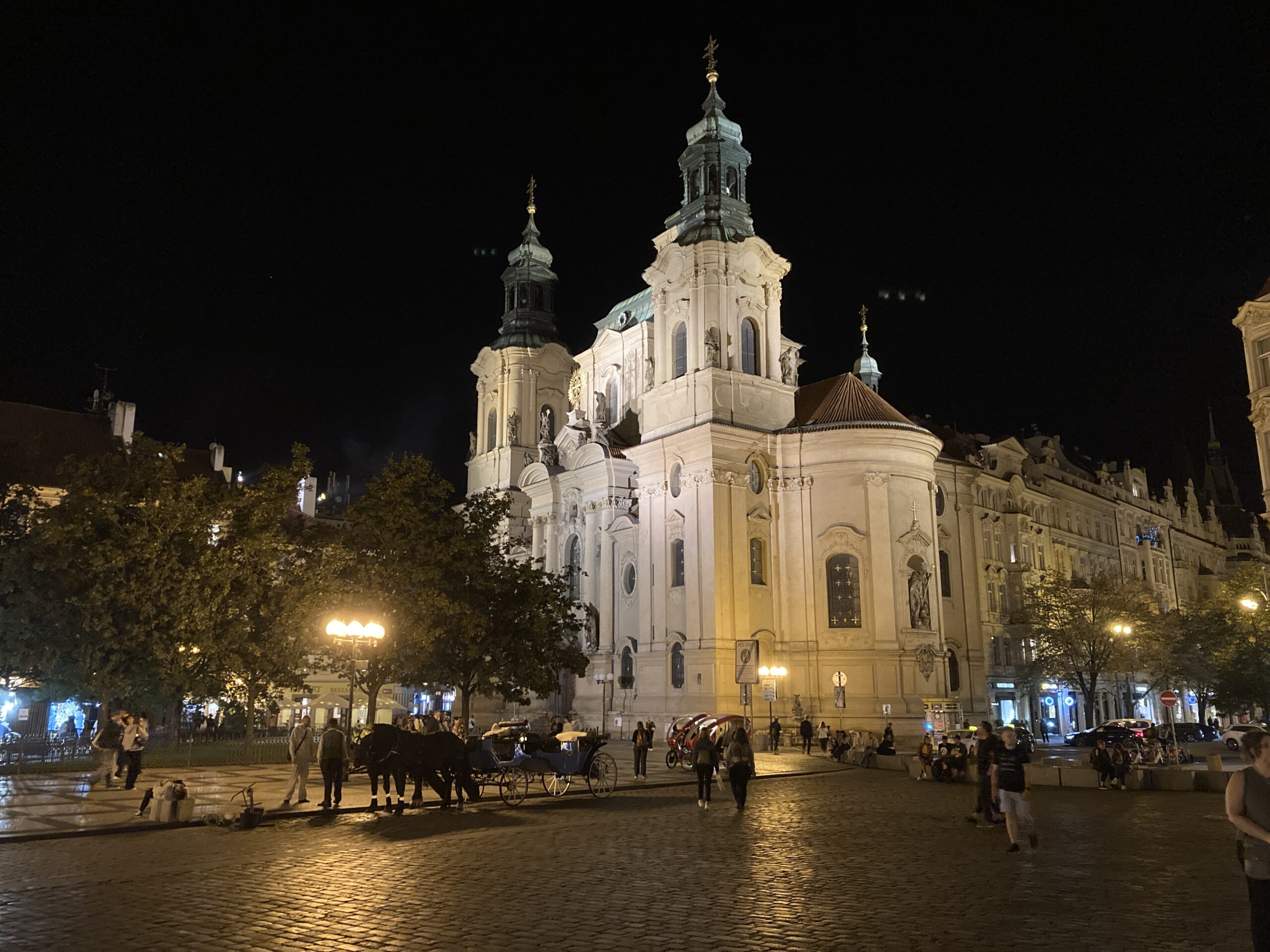 Beleuchtete St.-Nikolaus-Kirche am Altstädter Ring in Prag bei Nacht mit Passanten und Pferdekutschen.