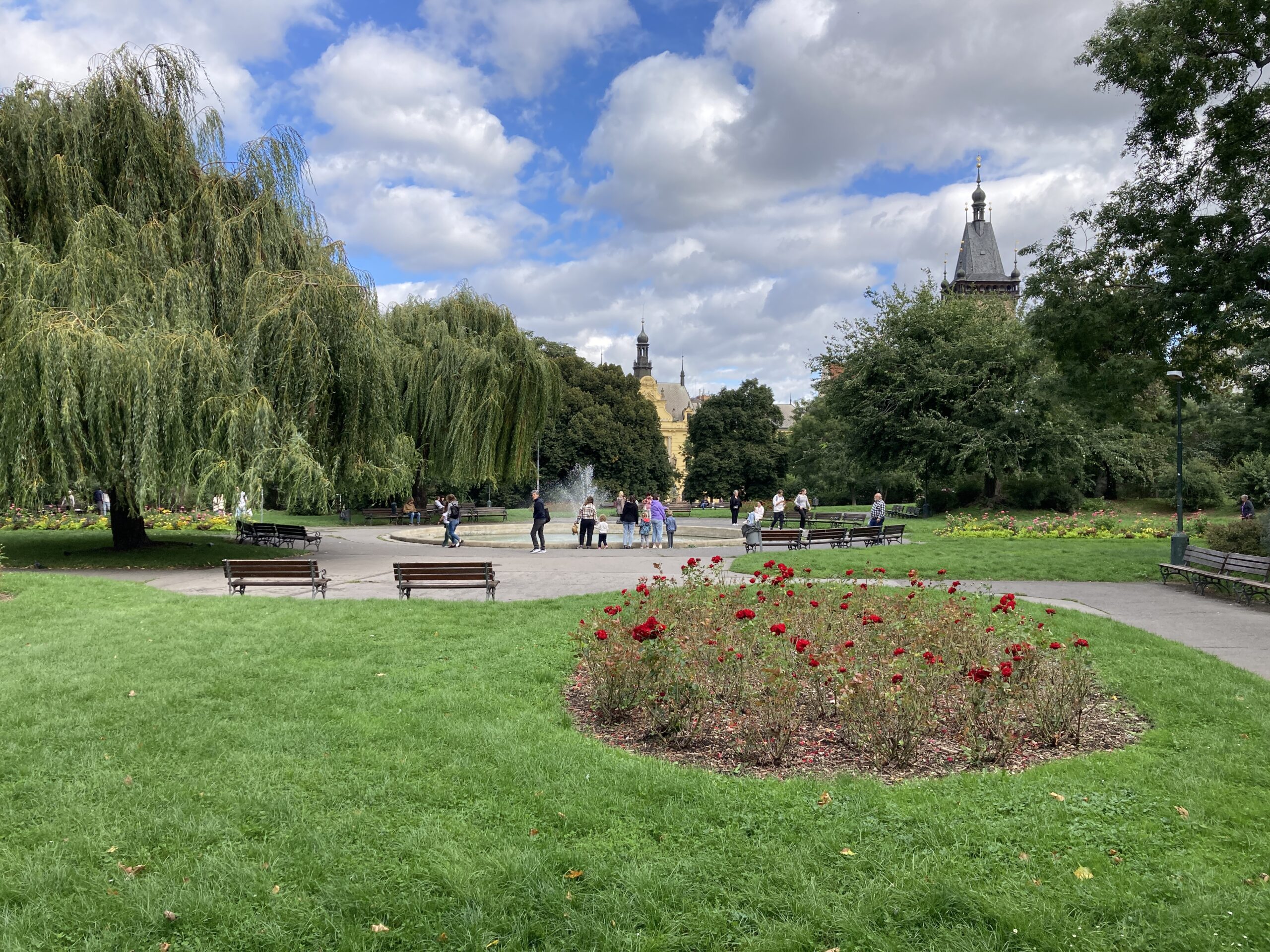 Grünanlage auf dem Karlsplatz in Prag mit Rasen, Rosenbeet, Bänken und großen Bäumen.
